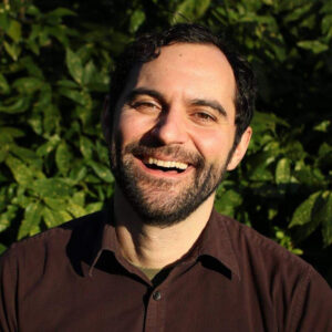 A man smiling for the camera in front of some bushes.