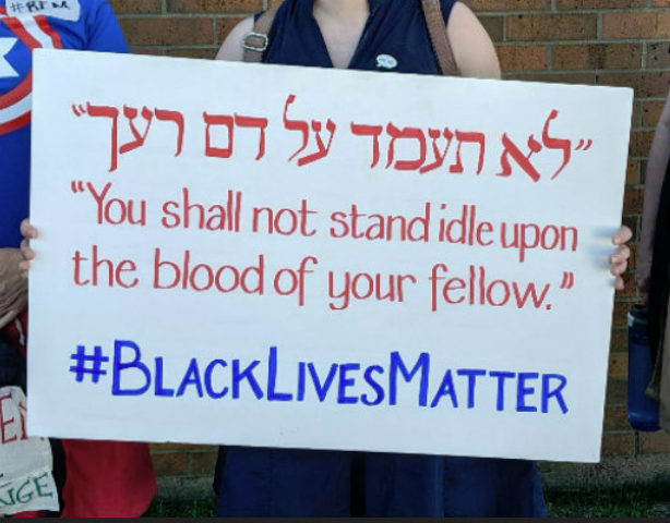 A woman holding up a sign that says " you shall not stand idle upon the blood of your fellow # blacklivesmatter ".