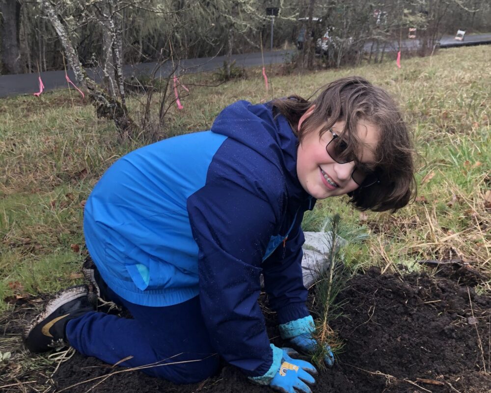 A girl in blue jacket kneeling on ground near trees.